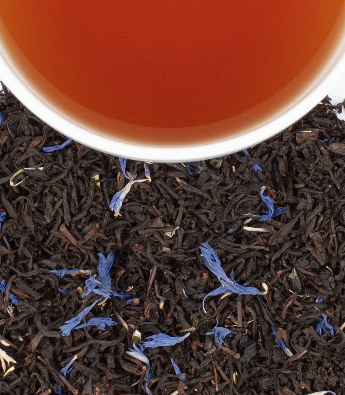 A cup of Celebration Tea sits atop loose black tea leaves and blue cornflower petals, showcasing its rich color.