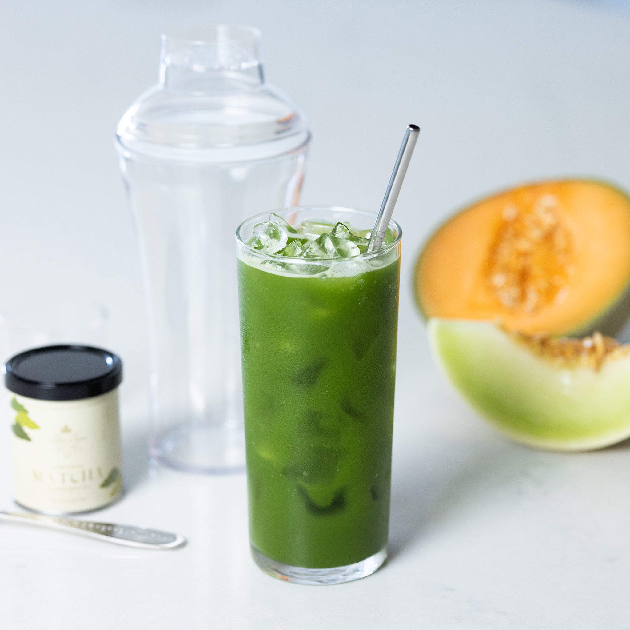 Iced matcha drink in a glass with ice, shaker, matcha powder, and melon slice on a light background.